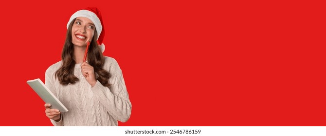 Christmas Wishes. Young European woman in Santa Claus hat happily writes her holiday wishlist in notebook and planning New Year, posing on red studio backdrop. Panorama, free space - Powered by Shutterstock