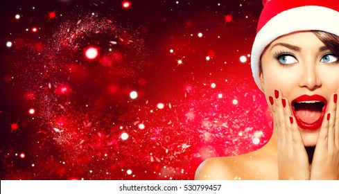 Christmas woman. Beauty model girl in Santa Claus hat with red lips and manicure looking right with a surprised expression. Closeup portrait over red holiday snow wide background copy space. Emotions.