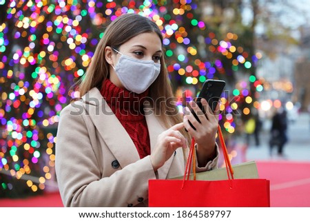 Christmas woman wearing face mask on street holding shopping bags and smart phone for online purchases with colorful christmas tree lights on background