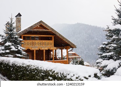 Christmas Wooden Mansion In Mountains On Snowfall Winter Day. Cozy Chalet On Ski Resort Near Pine Forest. Cottage Of Round Timber With Wooden Balcony. Fir-trees Covered With Snow. Chimneys Of Stone.