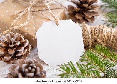 Christmas Wrapped Present With Paper Gift Tag On A White Table With Fir Tree Branches And Decorations Close Up. Rustic Winter Composition With Blank Gift Tag Mockup, Copy Space