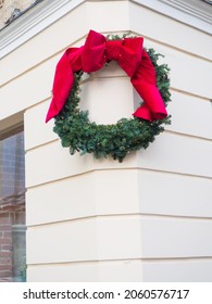 Christmas Wreath Decorated With Red Ribbon Bow Hanging On The Beige Wall. Close Up View Of Christmas Decorations Outside During The Festive Period In Lviv. Big Green Wreath On The Corner Of The House.