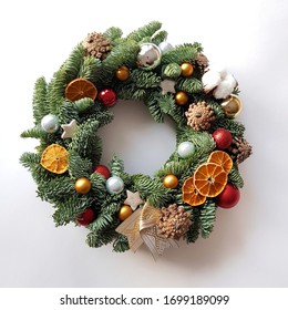 Christmas wreath lies on a white background. Decor elements are cones, glass balls, dried mugs of oranges, Christmas stars, cotton bolls. The basis of the wreath is spruce branches.
