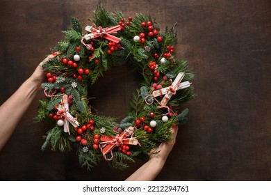 corona navideña de ramas naturales eververdes con bayas rojas en manos de mujer sobre fondo marrón de madera. Decoración festiva para vacaciones navideñas. Vista desde arriba.