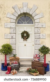 Christmas Wreath On An Imposing Door Of An English Country House, Suitcases And Gifts Line The Steps With Bay Trees In Pots On Each Side. Suitable For A Christmas Card Or Seasonal Magazine Cover.