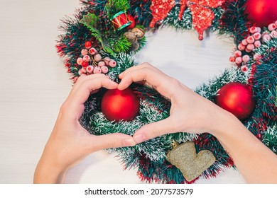 Christmas Wreath On Wooden Background. Women's Hands Show Heart Symbol On Background Of Christmas Wreath With Red Balls. Symbolic Sign Of Love With Hands With Christmas Decorations. Advent Family, DIY