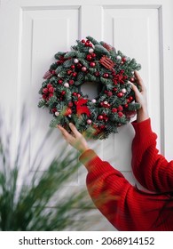 Christmas wreath with red balls and toys in female hands. Frontal view on a white wooden door. New year and christmas concept