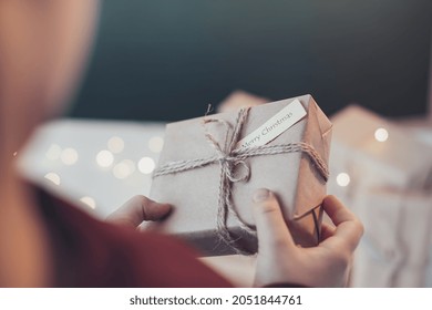 Christmas And Zero Waste, Environmentally Friendly Packaging.Children's Hands Holding Handmade Square Gift From Kraft Paper A Tag For Handmade Gifts On The Background Of Other Gifts. 