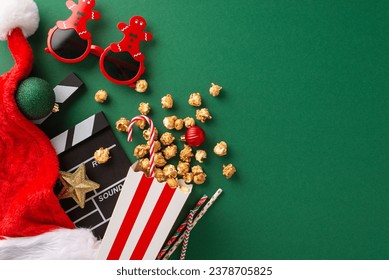 Christmas-themed premiere featuring thematic elements. Top-view of movie clapper, gingerbread man-shaped glasses, delectable popcorn, Santa's hat, ornaments, straws on green backdrop with ad space - Powered by Shutterstock