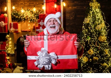Christmastime. Smiling Santa Claus with Christmas gift box in room decorated for Christmas. Winter family holiday. Bearded man in Santa hat with Christmas present. New Year eve. Christmas celebration.