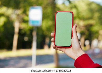 Chroma Key Phone In Red Case On Bus Stop Background, Female Arm Holding Green Screen Smartphone While Waiting Public Transport