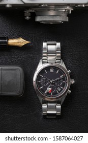 A Chrome Wristwatch For Man, Resting On Black Background. A Vintage Pen And A Ring And A Film Camera And Car Keys Are Visible Too In The Frame, Shot From Directly Above.