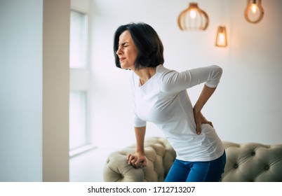 Chronic back pain. Adult woman is holding her lower back, while standing and suffering from unbearable pain. - Shutterstock ID 1712713207