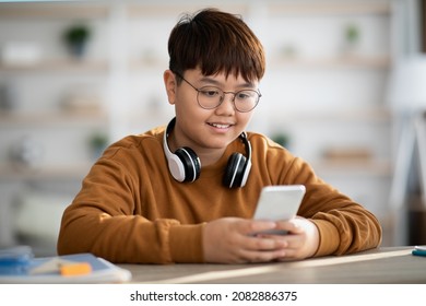 Chubby Asian Kid Boy In Casual Outfit Sitting At Table And Using Mobile Phone And Wireless Headset At Home, Playing Online Games Or Using Entertaining Mobile Application. Kids And Gadgets Concept