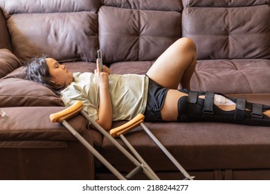 Chubby Asian Teen Girs With Injured Leg And Crutches Exting On Phone At Home, Lying On The Sofa