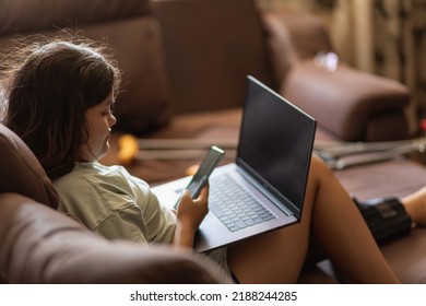 Chubby Asian Teen Girs With Injured Leg And Crutches Talking On The Phone And Laptop At Home, Sitting On The Sofa