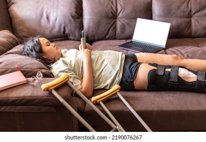 Chubby Asian Teen Girs With Injured Leg And Crutches Talking On The Phone And Laptop At Home, Lying On The Sofa