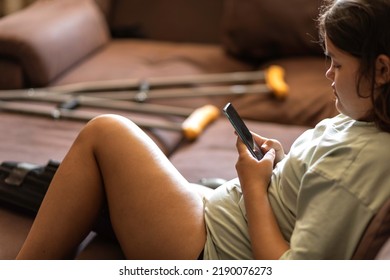 Chubby Asian Teen Girs With Injured Leg And Crutches Talking On The Phone At Home, Sitting On The Sofa