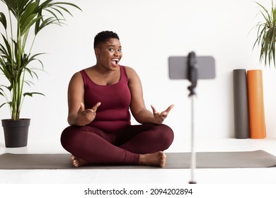 Chubby young african american woman in sportswear fitness coach having stream while exercising at white yoga studio, sitting on fitness mat, looking at smartphone set on tripod and gesturing - Powered by Shutterstock