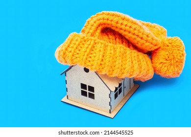Chunky Orange Pompom Beanie On Top Of A Toy House Model Isolated On A Blue Background. Home Heating Tips And Tricks Concept.