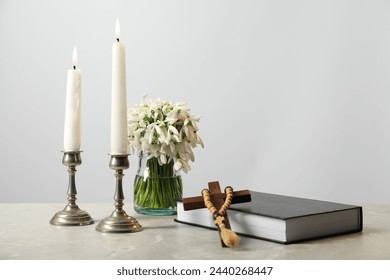 Church candles, wooden cross, rosary beads, Bible and flowers on marble table. Space for text