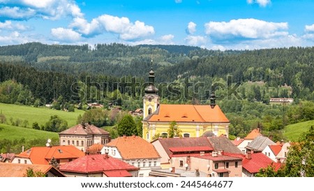 The Church of Saint Bartholomew in Pecka towers over quaint houses amidst lush green hills under a clear blue sky. Czechia