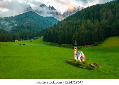 The Church of St. Johann is in a beautiful scenic location against the Geisler peaks covered with clouds in Santa Maddalena village, Val Di Funes, Italy. 