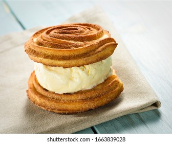 Churro Ice Cream Sandwich, Made With Homemade Churros And Vanilla Ice Cream. A Delicious Dessert Served On A Bege Napkin On A Blue Wooden Table.