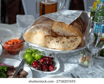 Ciabatta bread on the table in a restaurant. Table set with variety of dishes: loaf of bread, bowl of salsa, plate of grapes and cherries, glass of beer, plate of meat