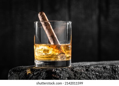 Cigar In A Glass Of Whisky With Ice On A Burnt Black Wood.