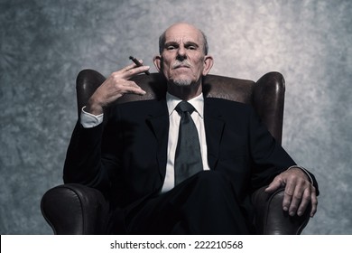 Cigar rauchend Senior Business-Mann mit grauem Bart mit dunklem Anzug und Krawatte. Sitzen auf Lederstuhl. Gegen graue Wand.