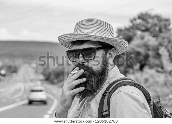 Cigarette before long journey. Traveler stylish\
hipster take brake.Man with beard and mustache in straw hat smoking\
cigarette, road background defocused. Smoking cigarette before long\
journey.