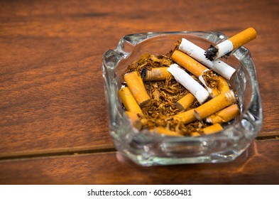 Cigarettes And Tobacco Lying Inside And Around Glass Ash Tray On Wooden Surface, Seen From Above, Anti Smoking Concept