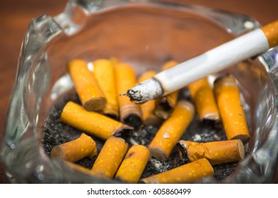 Cigarettes And Tobacco Lying Inside And Around Glass Ash Tray On Wooden Surface, Seen From Above, Anti Smoking Concept