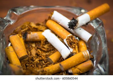 Cigarettes And Tobacco Lying Inside And Around Glass Ash Tray On Wooden Surface, Seen From Above, Anti Smoking Concept