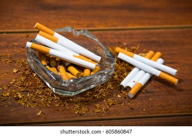 Cigarettes And Tobacco Lying Inside And Around Glass Ash Tray On Wooden Surface, Seen From Above, Anti Smoking Concept