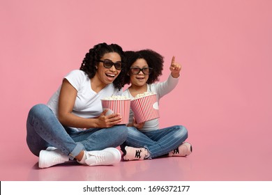 Cinema Fun. Cheerful African Mom And Daughter Watching Movie In 3d Glasses And Eating Popcorn, Sitting On Floor In Studio Over Pink Background