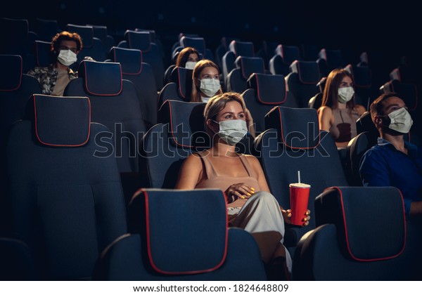 Cinema in quarantine. Coronavirus pandemic\
safety rules, social distance during movie watching. Men, women in\
protective face mask sitting in a rows of auditorium. Leisure time,\
youth culture concept.