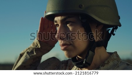 Cinematic close up shot of young serious determined responsible female patriot soldier in camouflage military uniform and helmet is saluting on horizon on fields at sunset.