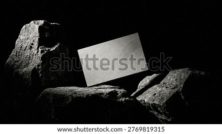 A cinematic composition featuring a light grey business card levitating between raw, dark rocks. The black background and side lighting emphasize the mineral textures, creating a minimalist and sophisticated luxury atmosphere.