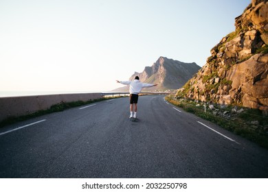 Cinematic Epic Photo Of Young Man In Grey Hoodie Ride Longboard On Empty Road In Beautiful Mountain Landscape. Wanderlust Travel Concept. Millennial Generation Z Adventure In Summer. Sunset Skating