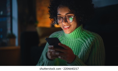 Cinematic Portrait of Black Female with Curly Hair Sitting on the Floor at Home and Using Smartphone. Diverse Woman Receives Good News and Chatting with Friends Via Social Media.