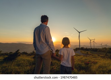 Kinematische Aufnahme eines sorgenfreien Jungvateringenieurs, der seine Tochter für die Hand hält und bei Sonnenuntergang auf das Feld der Windmühle schaut. Konzept der erneuerbaren Energien, Liebe für die Natur, Familie, Strom, umweltfreundliche Energie, Zukunft