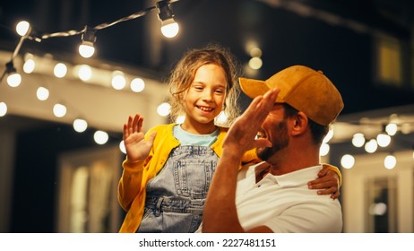 Cinematic Shot of a Father Holding His Small Daughter in His Arms, Helping Her to Fix a Lightbulb in a Backyard Lights Installation. Father and Daughter High Five and Celebrate Successful Repair.