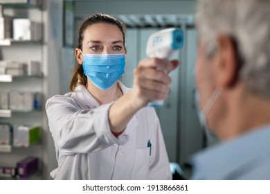 Cinematic Shot Of Female Pharmacist With Medical Mask Is Measuring Temperature With Infrared Thermometer Gun To Male Patient In Pharmacy. Concept Of Covid-19, Protection, Virus Transmission, Safety.