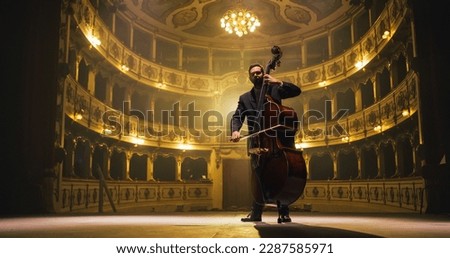 Cinematic shot of Male Cellist Playing Cello Solo on an Empty Classic Theatre Stage with Dramatic Lighting. Professional Musician Rehearsing Before the Start of a Big Show with Orchestra