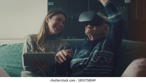 Cinematic shot of senior grandfather with vr glasses and granddaughter with tablet are having fun to use augmented reality technology together for family entertainment while sitting on sofa at home. - Powered by Shutterstock