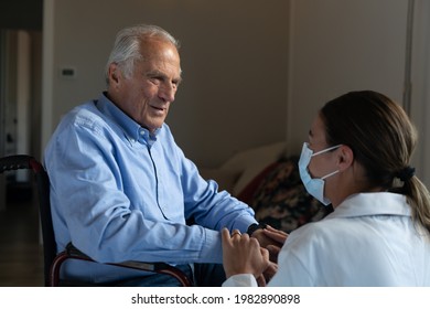 Cinematic Shot Of Social Worker With Medical Mask Stroking Hands Of Senior Man As Sign Of Care And Respect In Wheelchair At Home. Concept Of Healthcare, Protection, Assistance, Caregiver, Covid-19.