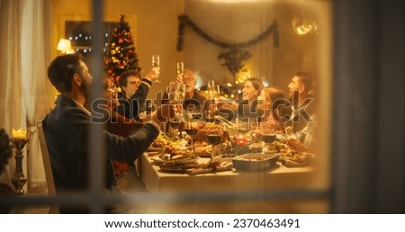 Cinematic Shot Through a Snowy House Window: Christmas Dinner Together with Parents, Children and Friends at Home. Multicultural Family Raising Champagne Glasses and Toasting, Celebrating Holidays