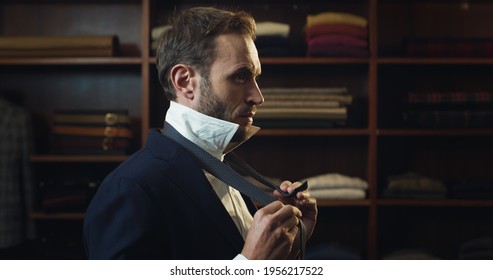 Cinematic Shot Of Young Businessman Is Tying A Tie While Trying High Quality Tailor Made Suit By Designer In Luxury Tailoring Atelier. Concept Of Fashion, Handmade, Hand Craft, Couturier, Business.